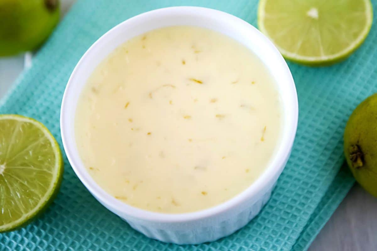 A white ramekin filled with a creamy and textured key lime pie filling is placed on a teal textured cloth. Sliced halves of fresh green limes surround the ramekin. The filling appears smooth with small pieces of zest visible throughout.