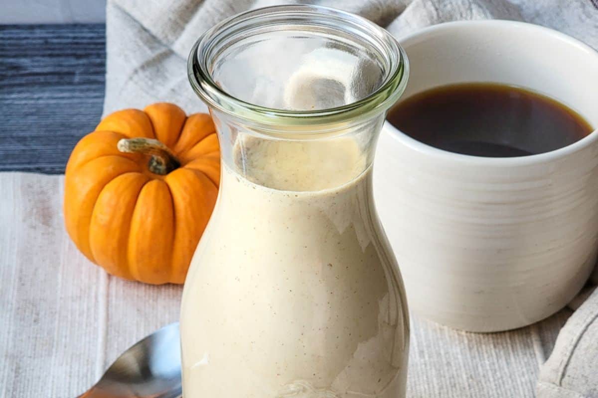 A glass bottle filled with creamy, light beige liquid sits on a white cloth next to a small orange pumpkin. A white mug filled with dark coffee is also visible in the background along with a metal spoon lying on the cloth.