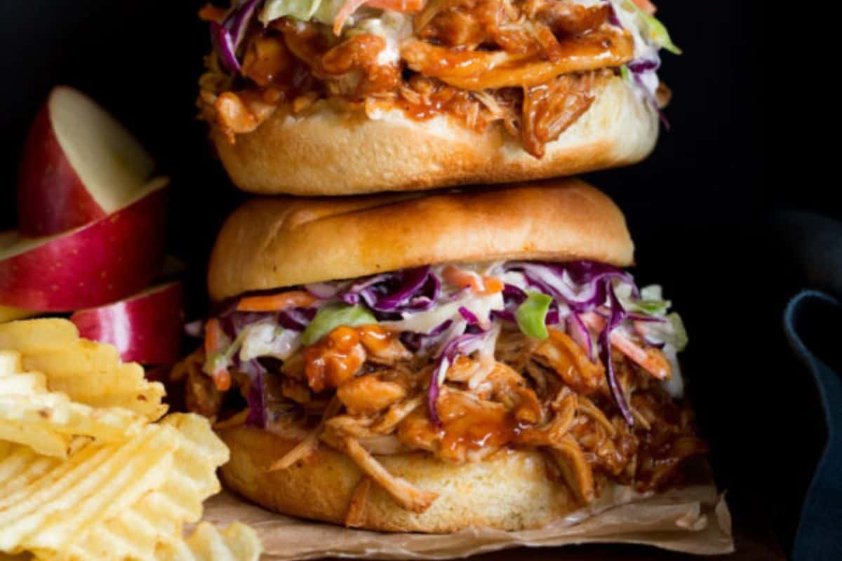 A close-up of two stacked barbecue pulled pork sandwiches with coleslaw and sauce, served on buns. On the left side of the sandwiches, there are sliced red apple wedges, and on the bottom left corner, there are ripple-cut potato chips.