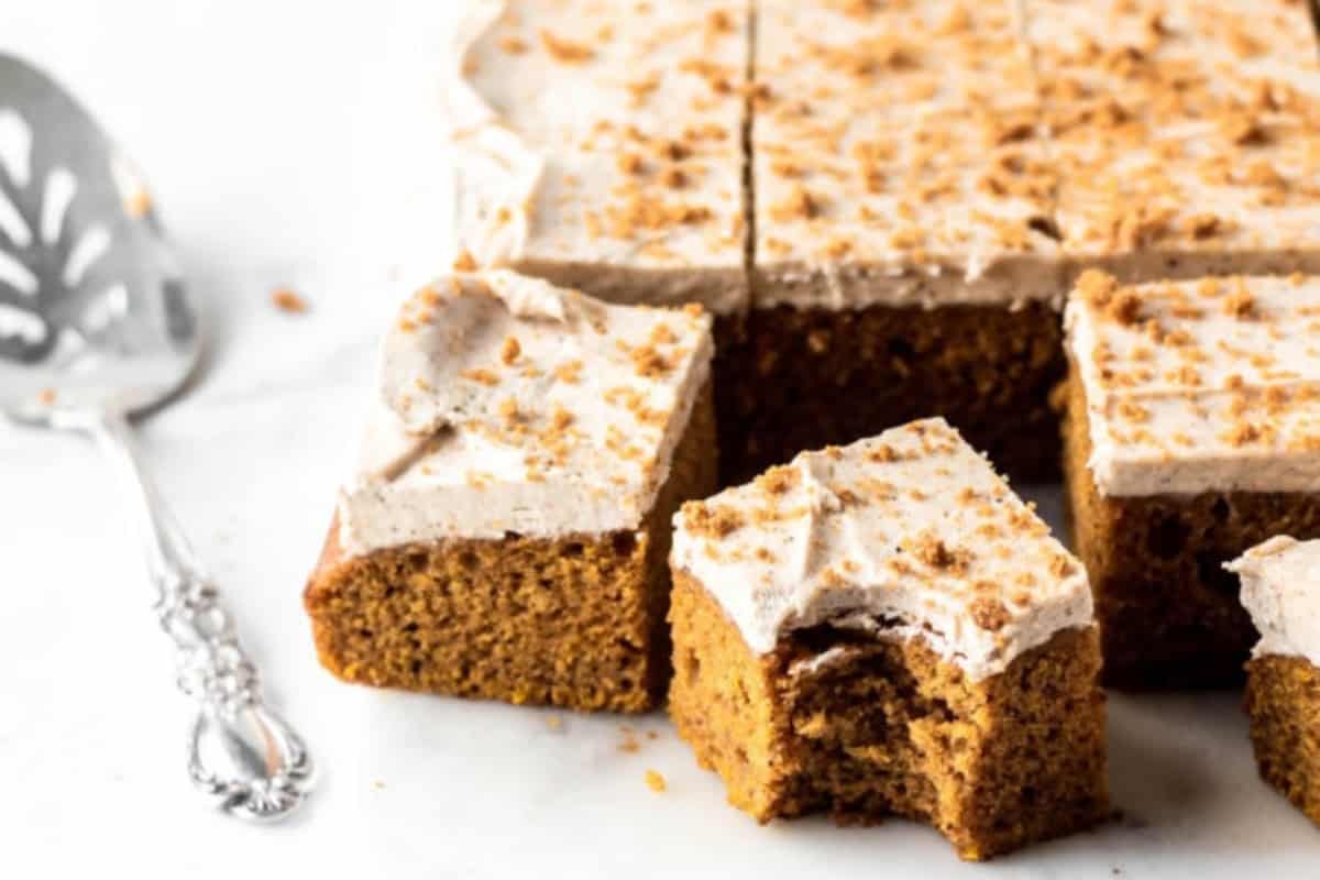 A tray of spiced sheet cake with white frosting, one piece on the side with a bite taken out. A silver serving spatula is next to the cake on a white surface.