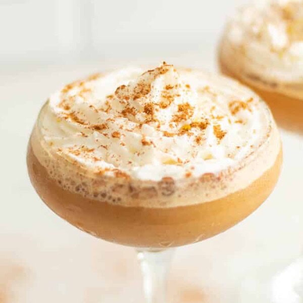 Two cocktail glasses filled with a creamy, frothy brown beverage topped with whipped cream and a sprinkle of cinnamon. The drink appears rich and inviting, likely flavored with spices, and the background is softly blurred, keeping the focus on the beverages.