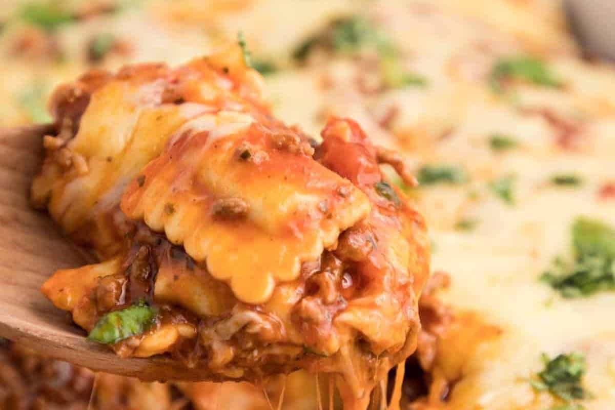 A close-up shot of a cheesy ravioli casserole being served. The ravioli are covered in melted cheese, tomato sauce, and pieces of minced meat, garnished with green herbs. The image shows the gooey, savory dish in detail with stretchy, melted cheese pulling away.