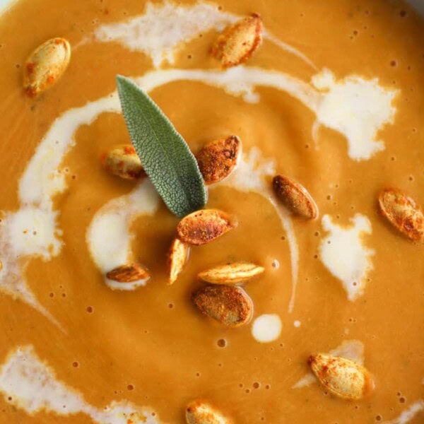 A bowl of creamy soup topped with roasted seeds, a drizzle of cream, and a sage leaf.