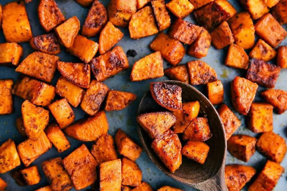 Roasted sweet potato cubes seasoned with spices, spread out on a baking sheet. Some cubes are scooped up with a wooden spoon.