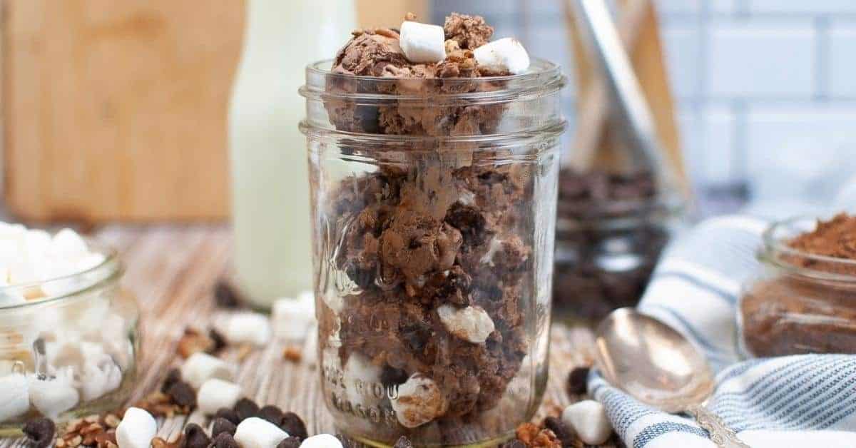 A glass jar filled with no-churn rocky road ice cream, topped with mini marshmallows, is surrounded by scattered marshmallows, chocolate chunks, and nuts. In the background, there are a bottle of milk and a spoon on a striped cloth.