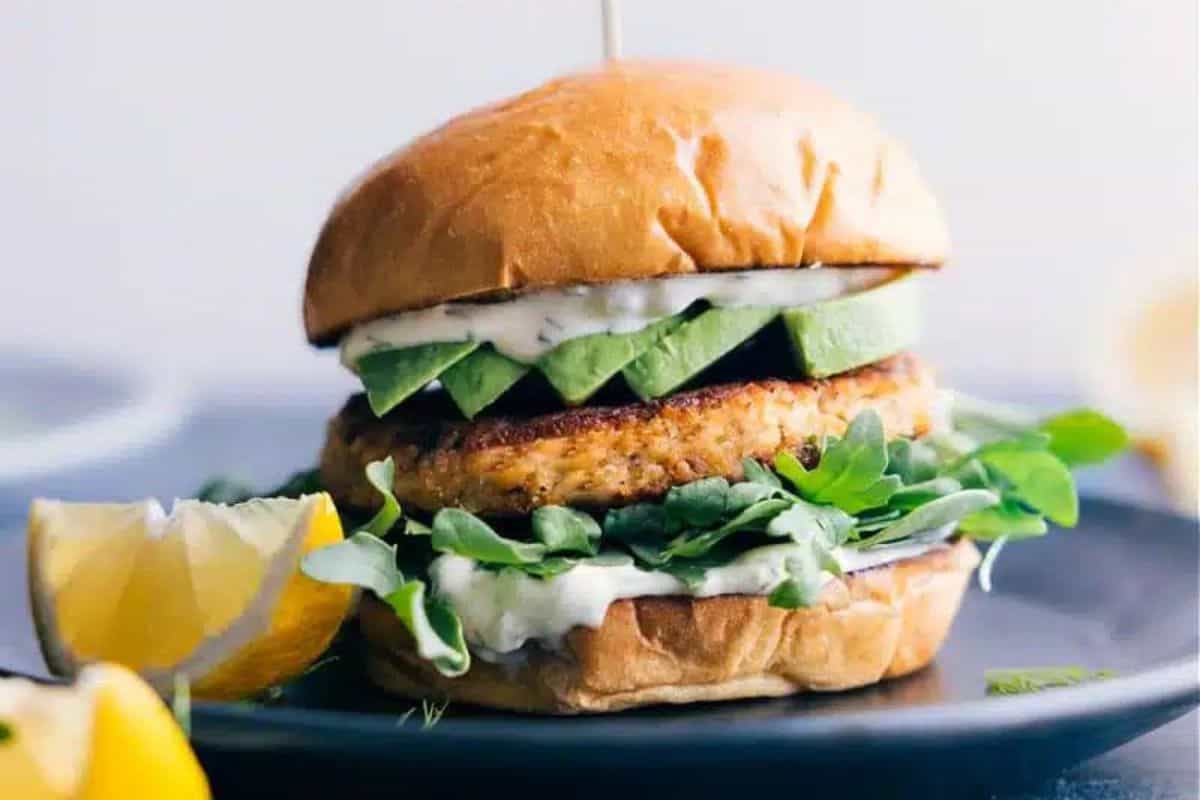 A close-up of a gourmet burger on a black plate. The burger features a golden-brown patty, fresh arugula, sliced avocado, and a dollop of creamy white sauce, all inside a toasted bun. A lemon wedge is placed next to the burger on the plate.