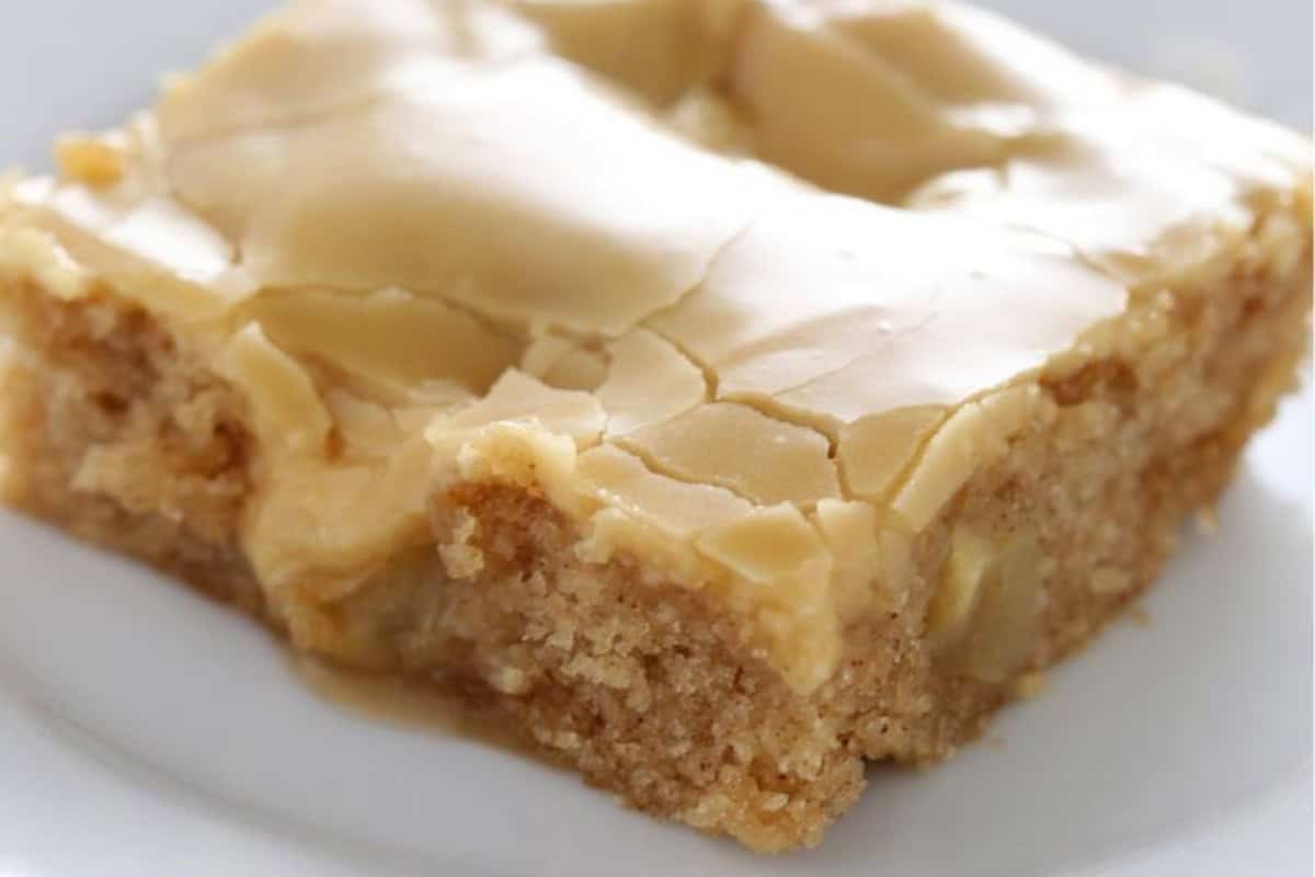 A close-up of a square slice of apple blondie bar on a white plate. The blondie is topped with a smooth layer of caramel glaze that is slightly cracked and has a moist, dense texture with visible apple chunks.