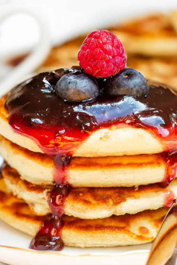 A stack of pancakes topped with berry sauce, a raspberry, and two blueberries sits on a floral plate with a fork. More pancakes are on a plate in the background.