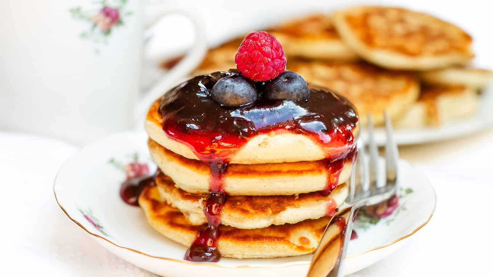 A stack of pancakes topped with blueberry and raspberry jam, garnished with fresh blueberries and a raspberry. The pancakes are on a white plate with a floral design, and a fork is resting on the side. A cup and a plate of more pancakes are blurred in the background.