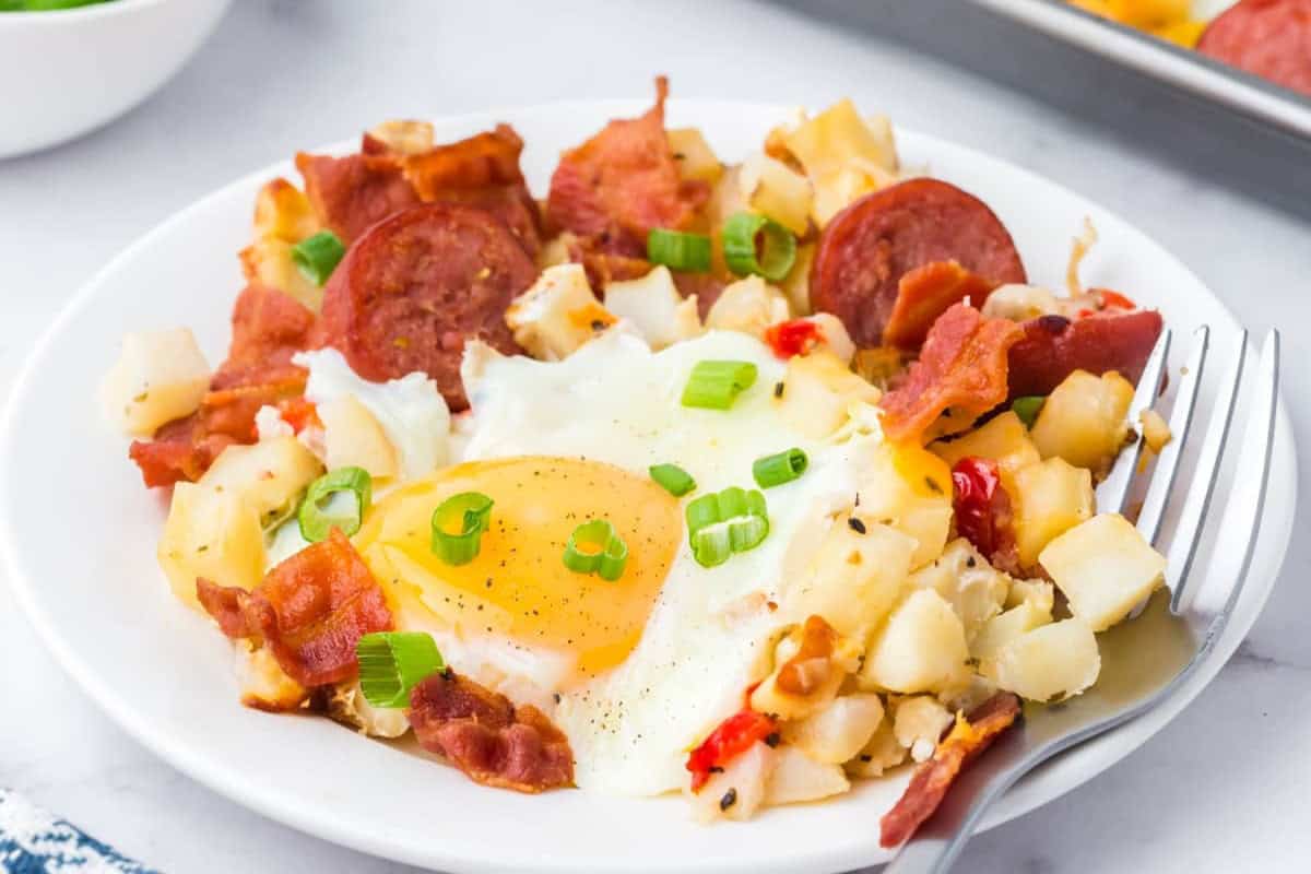 A plate of breakfast hash featuring a sunny-side-up egg surrounded by diced potatoes, crispy bacon strips, slices of sausage, chopped green onions, and small pieces of red bell pepper. A fork rests on the side of the plate.