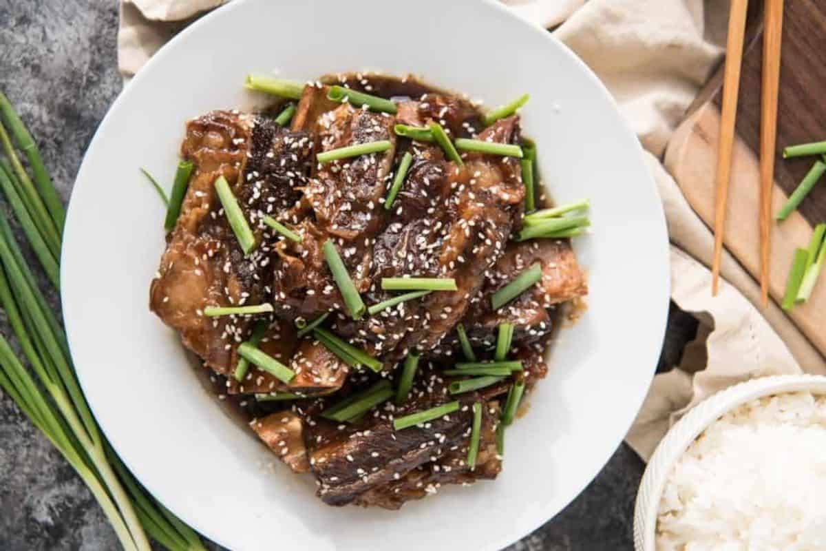 A white plate filled with braised beef short ribs garnished with green onions and sesame seeds. The dish is placed on a dark textured surface with chopsticks and a bowl of white rice nearby. Some green onions are also visible on the left side of the plate.