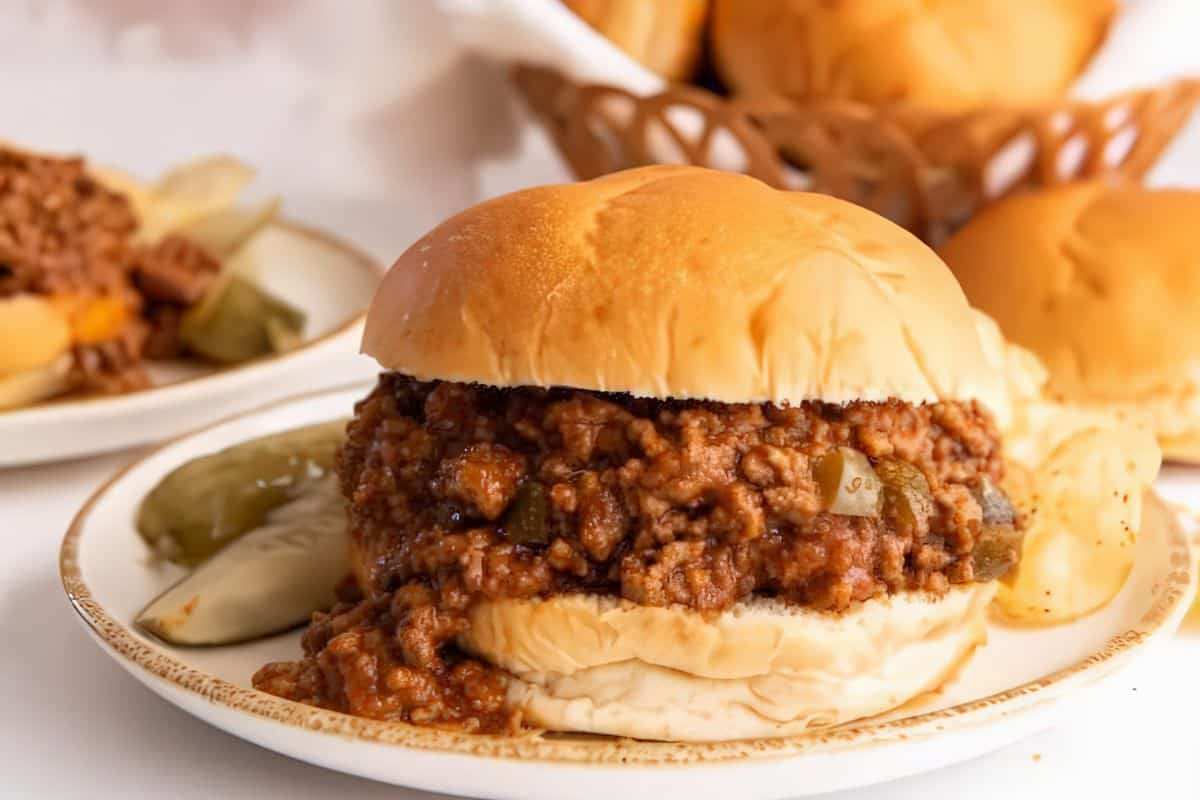 A sloppy joe sandwich on a white plate with pickle slices and potato chips, with a basket of buns in the background.