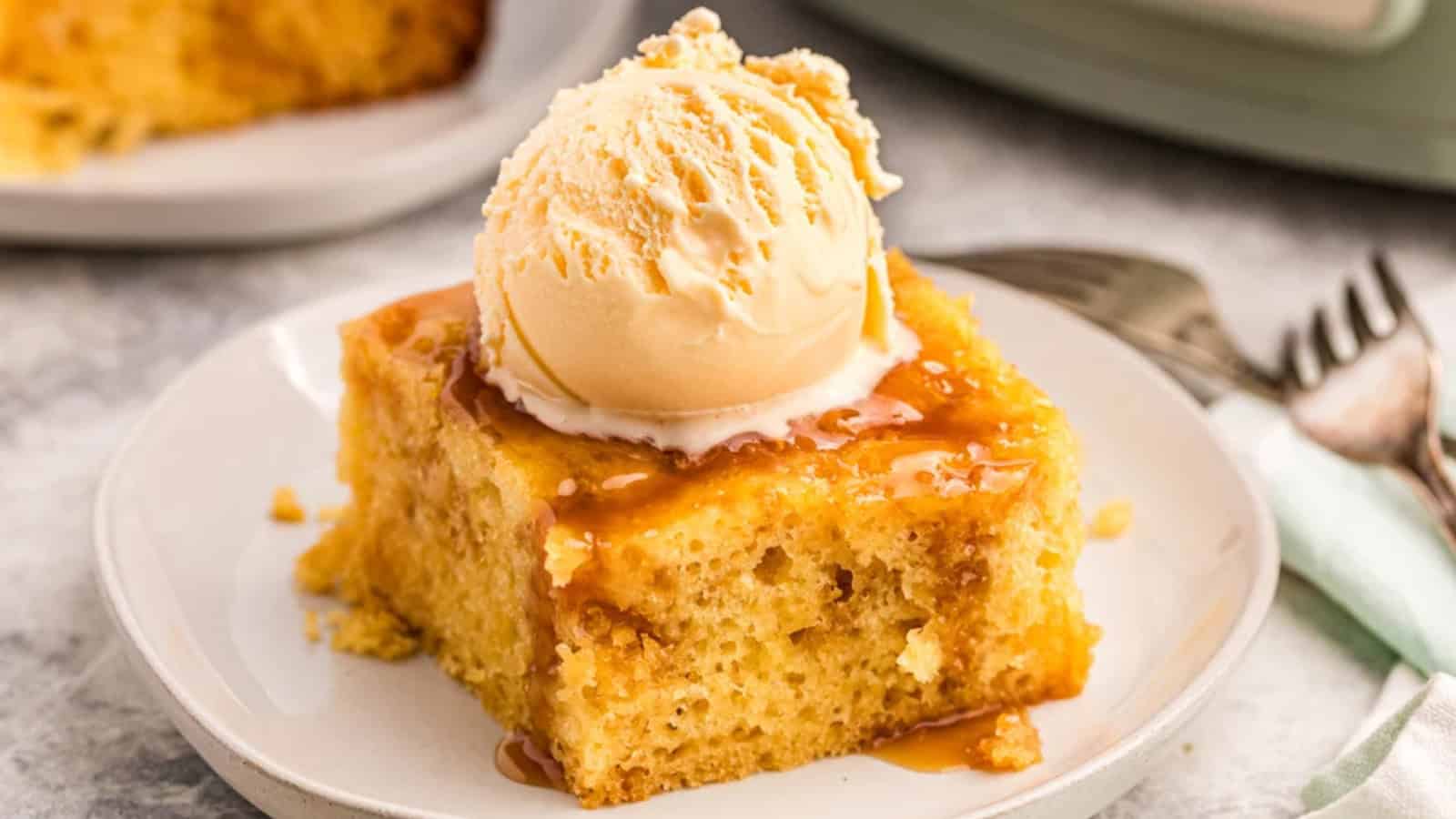 A slice of moist cake topped with a scoop of vanilla ice cream and drizzled with caramel sauce sits on a white plate. Forks and a napkin are beside it on a light-colored surface.