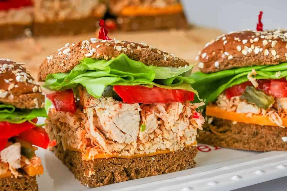 Close-up of a hearty sandwich made with whole grain bread, filled with shredded chicken, cheddar cheese, lettuce, tomato slices, and pickles. The sandwich is garnished with a red toothpick on top, and a blurred background showcases other similar sandwiches.
