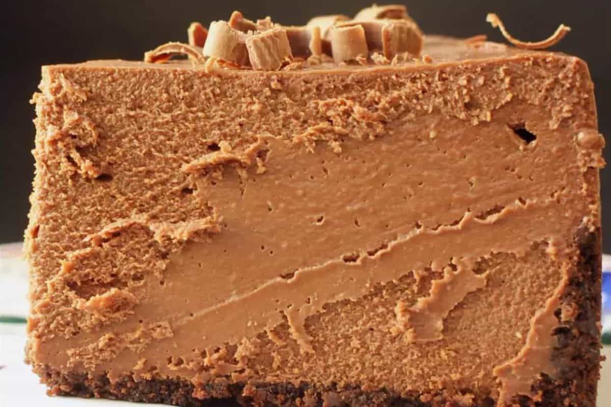 Close-up of a decadent chocolate cheesecake slice with a creamy texture and a rich, thick chocolate filling. The top is adorned with chocolate shavings, and the base is a dark chocolate crust.