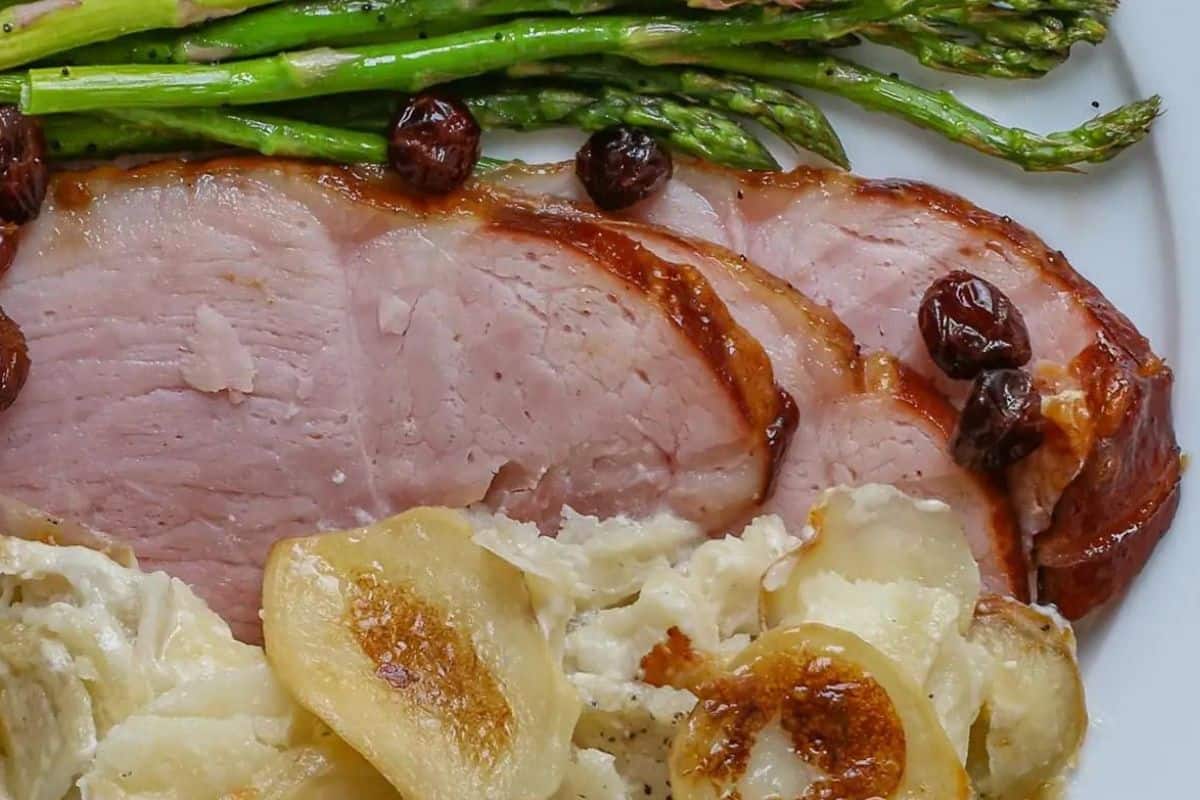 A close-up of a plate featuring slices of glazed ham, a few roasted cherries, sautéed asparagus spears, and a serving of creamy scalloped potatoes. The dish is presented on a white plate, highlighting the contrasting colors and textures.