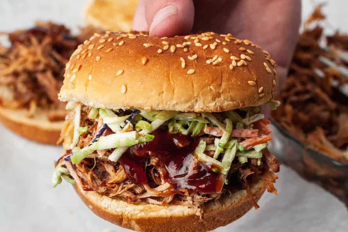 A close-up of a hand holding a pulled pork sandwich with a sesame seed bun. The sandwich is filled with shredded barbecue pork, coleslaw, and barbecue sauce. More pulled pork sandwiches are blurred in the background on a white surface.