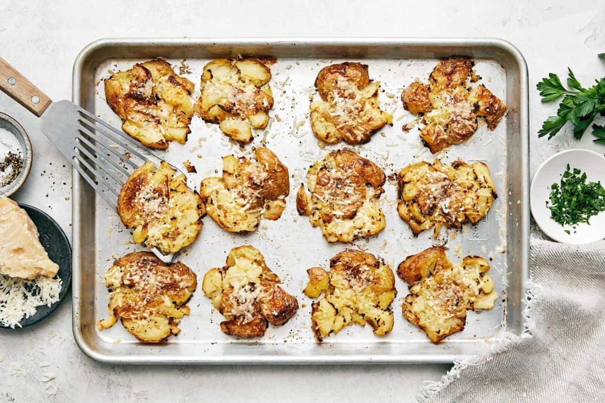 A baking sheet with 12 smashed potatoes, garnished with melted cheese and herbs. A metal spatula is lifting one potato. Surrounding items include a block of cheese with a grater, a bowl of chopped herbs, and fresh parsley on a light-colored surface.