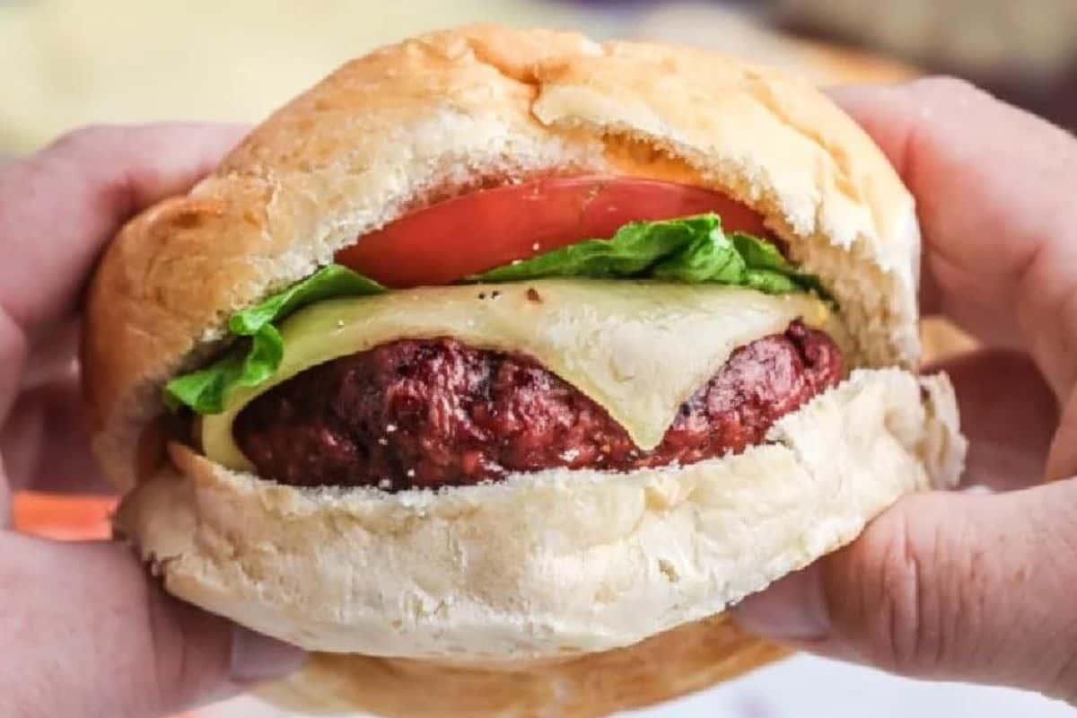 A close-up of someone holding a freshly made burger. The burger has a soft bun, a juicy patty, a slice of cheese, a piece of lettuce, and a slice of tomato. The hands are positioned to show the burger's layers clearly.
