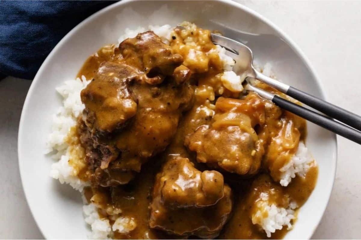 A white plate filled with steamed white rice topped with chunks of meat and gravy. The dish is served with black chopsticks and a fork placed on the side. The meal looks savory, and the gravy appears to be rich and thick. A blue napkin is partially visible to the left.