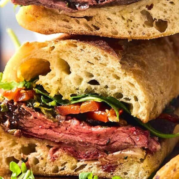 Close-up of a steak sandwich with arugula, roasted red peppers, and crusty bread. Some fries are partially visible in the background.