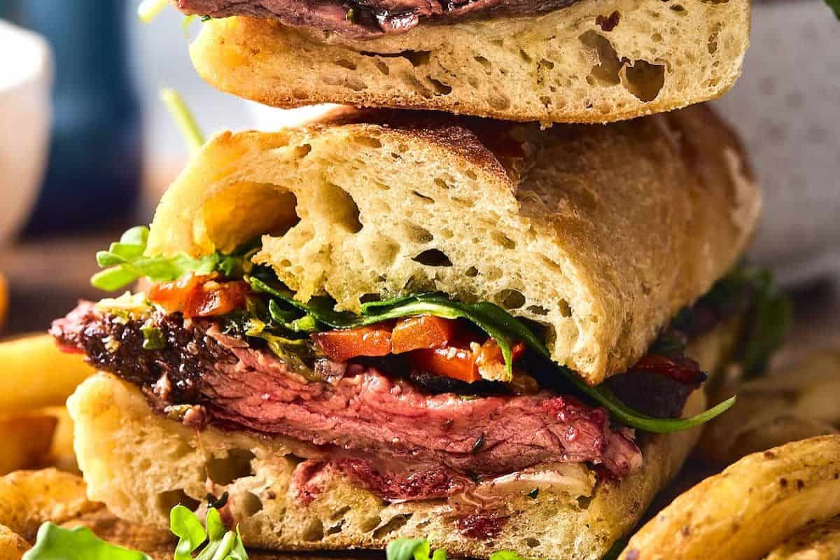 Close-up of a steak sandwich with arugula, roasted red peppers, and crusty bread. Some fries are partially visible in the background.