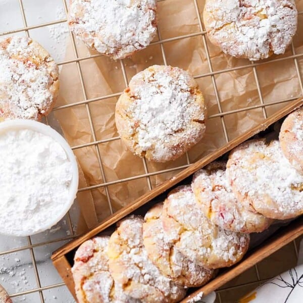 A cooling rack and tray filled with crinkle cookies dusted in powdered sugar; a bowl of powdered sugar nearby.