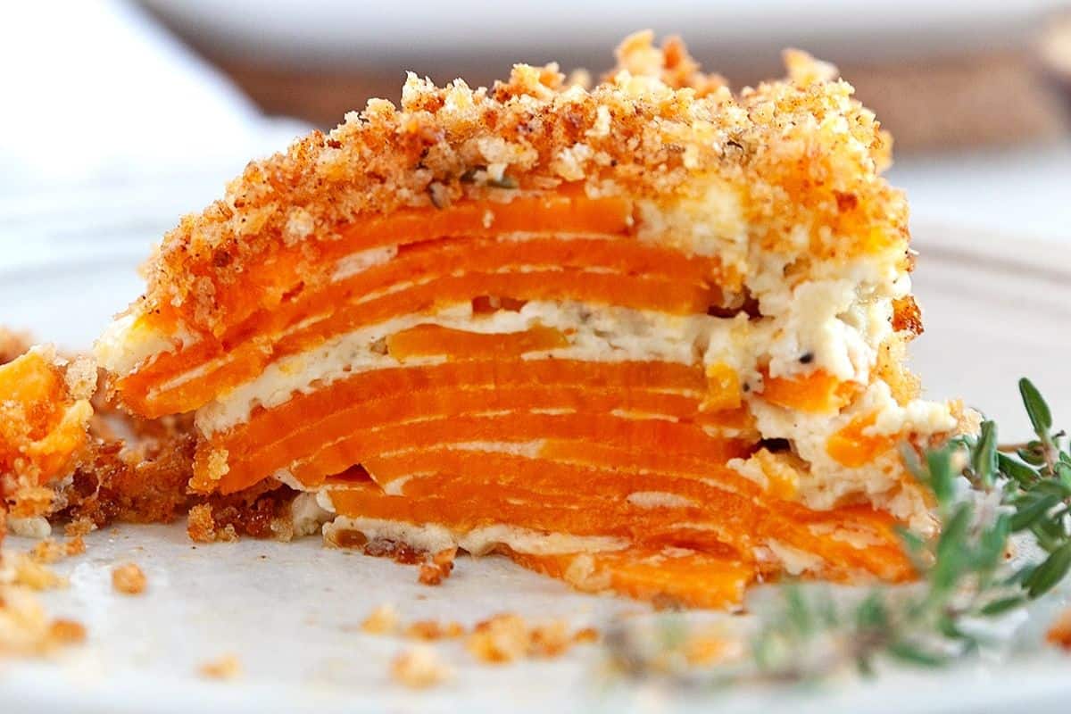 Close-up of a slice of sweet potato gratin. The layered sweet potatoes are covered with a crispy, golden breadcrumb topping, and creamy white cheese or sauce is visible between the layers. The dish rests on a white plate with a sprig of fresh herbs nearby.