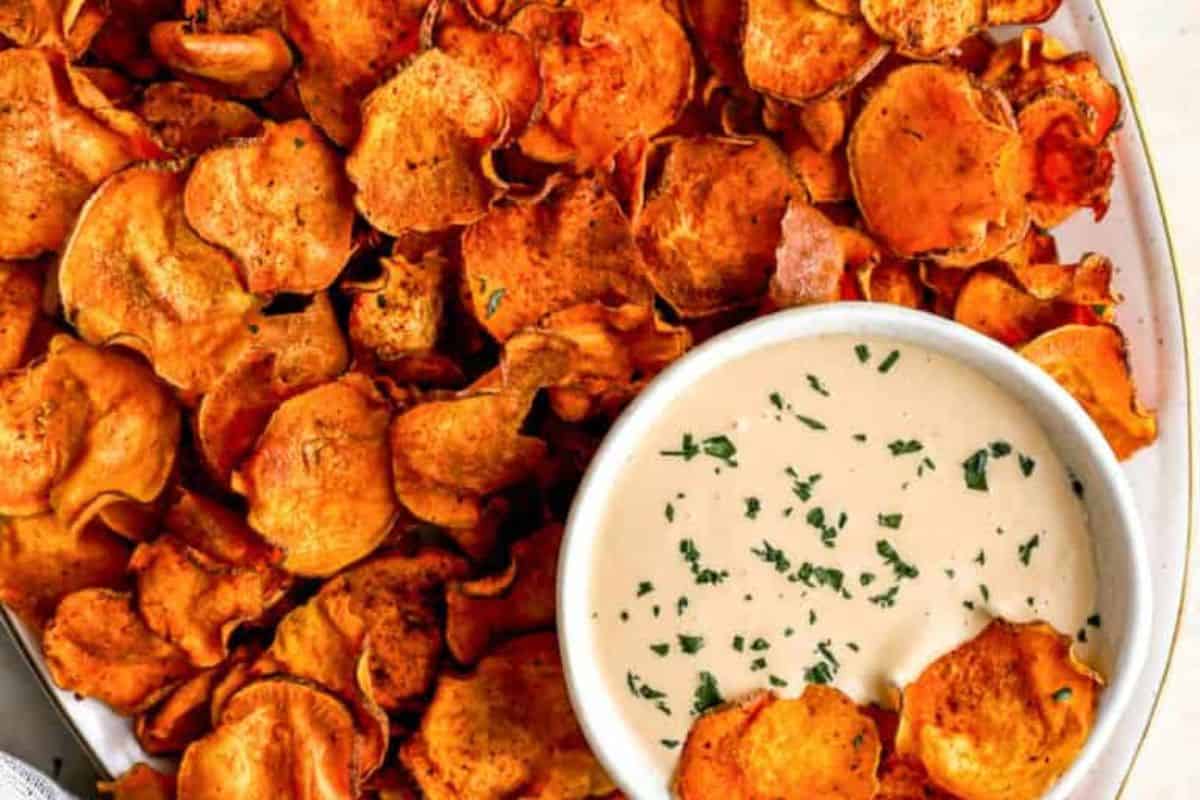 A plate of crispy sweet potato chips surrounds a small white bowl filled with creamy dipping sauce, garnished with chopped herbs. The golden-orange chips are slightly curled and look seasoned, contrasting with the smooth, light-colored dip.