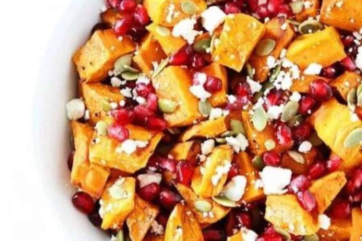 A close-up of a colorful salad featuring roasted sweet potatoes, pomegranate seeds, crumbled feta cheese, and pumpkin seeds, all mixed together in a white bowl.