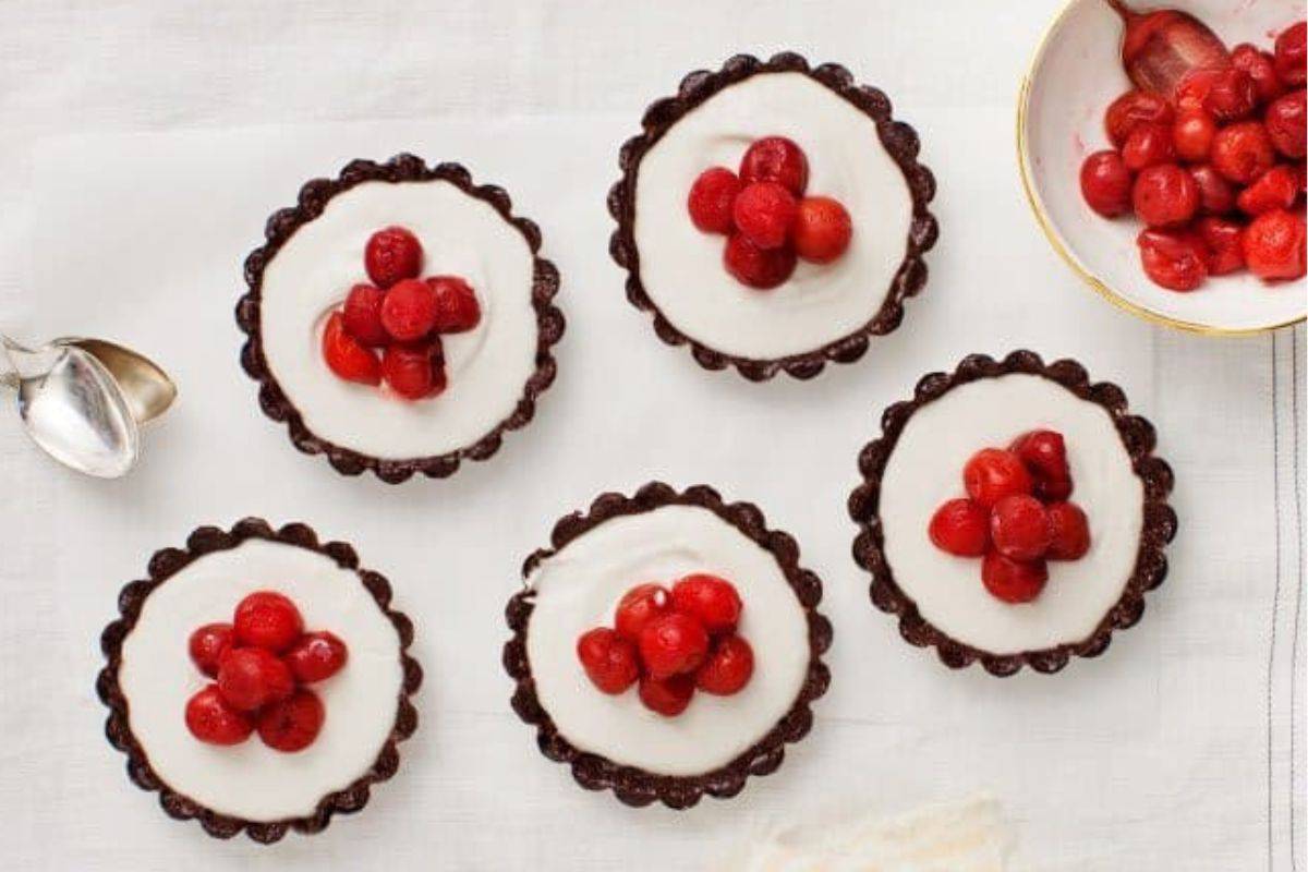 Five chocolate tarts with whipped cream filling and topped with red cherries are neatly arranged on a white surface. An additional bowl of red cherries and a silver spoon rest next to the tarts.