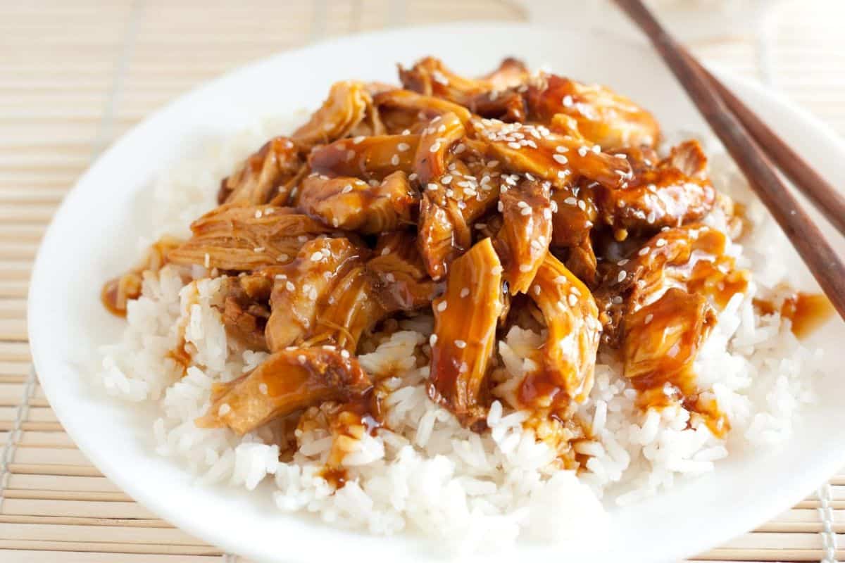 A plate of white rice topped with shredded teriyaki chicken, garnished with sesame seeds. A pair of wooden chopsticks rests on the edge of the plate. The background features a light-colored bamboo mat.