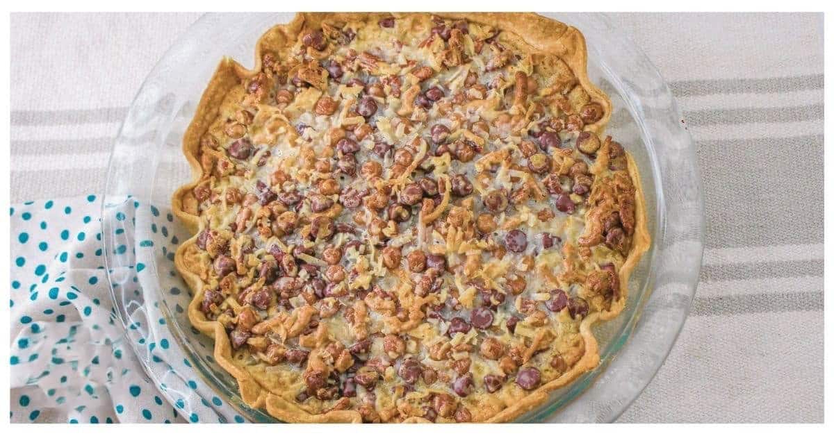A baked pie with a golden-brown crust filled with mixed nuts, caramel, and raisins is placed on a glass pie plate. The pie is set on a table covered with a gray and white striped tablecloth, alongside a white napkin with blue polka dots.