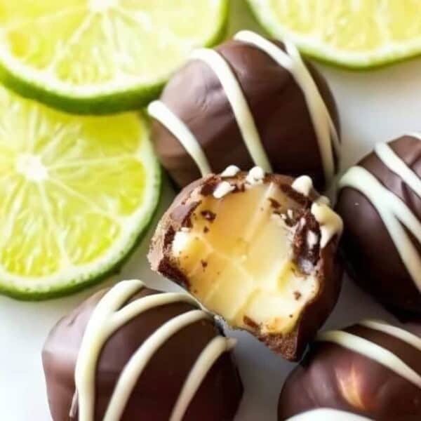 The image shows several dark chocolate truffles with white chocolate drizzle placed next to sliced key limes on a white surface. One truffle has a bite taken out, revealing a creamy interior.