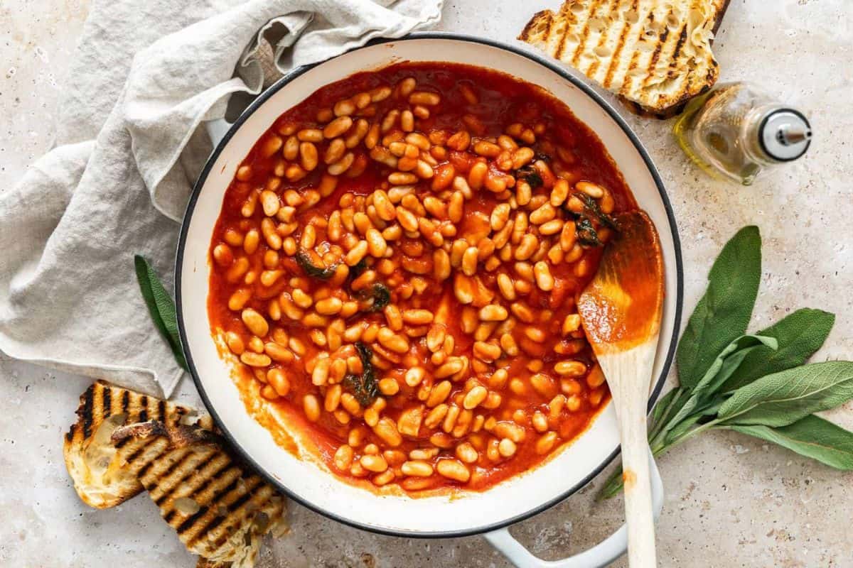 A pot of baked beans in a tomato sauce with a wooden spoon resting on the side. Grilled bread slices and fresh sage leaves are placed around the pot. A folded light-colored cloth is seen in the background. There is also a bottle of oil on the side.