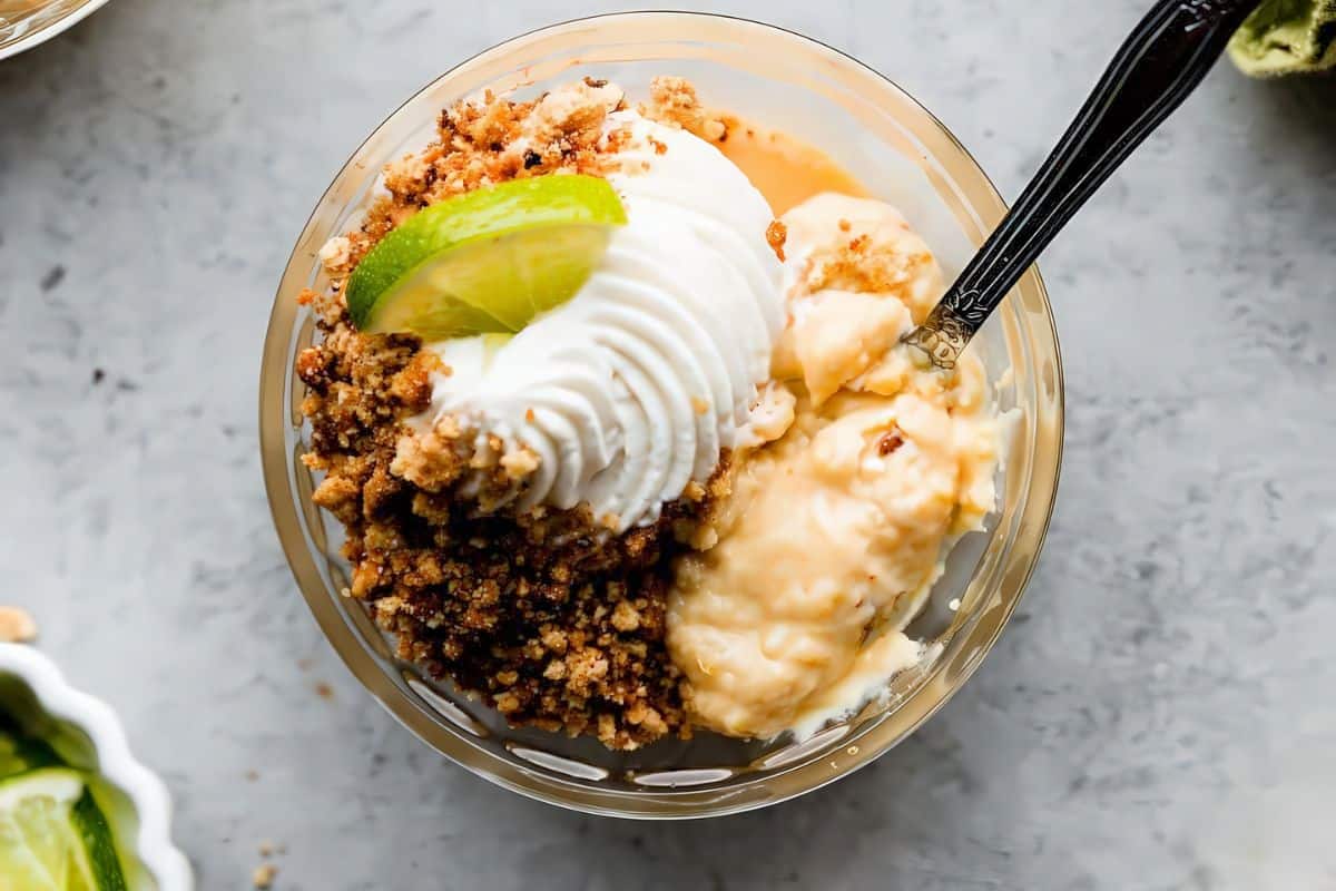 A glass bowl filled with creamy dessert topped with crumbled graham crackers, a dollop of whipped cream, and a slice of lime. A spoon is placed in the bowl. The bowl rests on a gray surface.