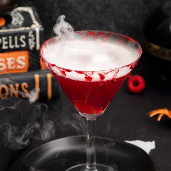 A red cocktail in a martini glass with smoky effect and bloody rim, surrounded by cocktail recipes, books, Chambord bottle, and spider.