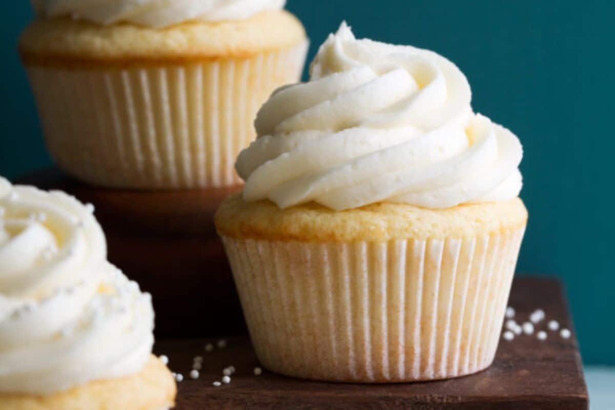 A close-up of three vanilla cupcakes with white frosting on a wooden surface. The cupcakes are neatly wrapped in white paper liners, and small white sprinkles are scattered in the background, with a teal-colored backdrop behind them.