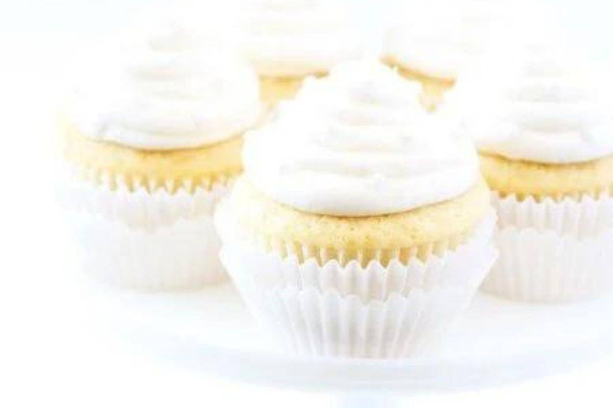 Five vanilla cupcakes with a swirl of white frosting on top, arranged closely together on a white surface. The cupcakes have white paper liners, creating a soft, monochromatic aesthetic.