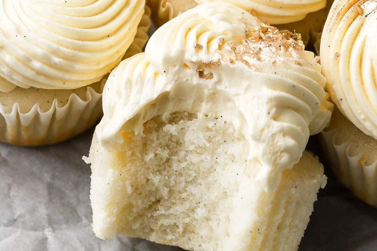 Close-up of vanilla cupcakes with swirls of creamy white frosting on top. The front cupcake has a large bite taken out of it, revealing its fluffy texture. The tops of the cupcakes are slightly sprinkled with flakes.