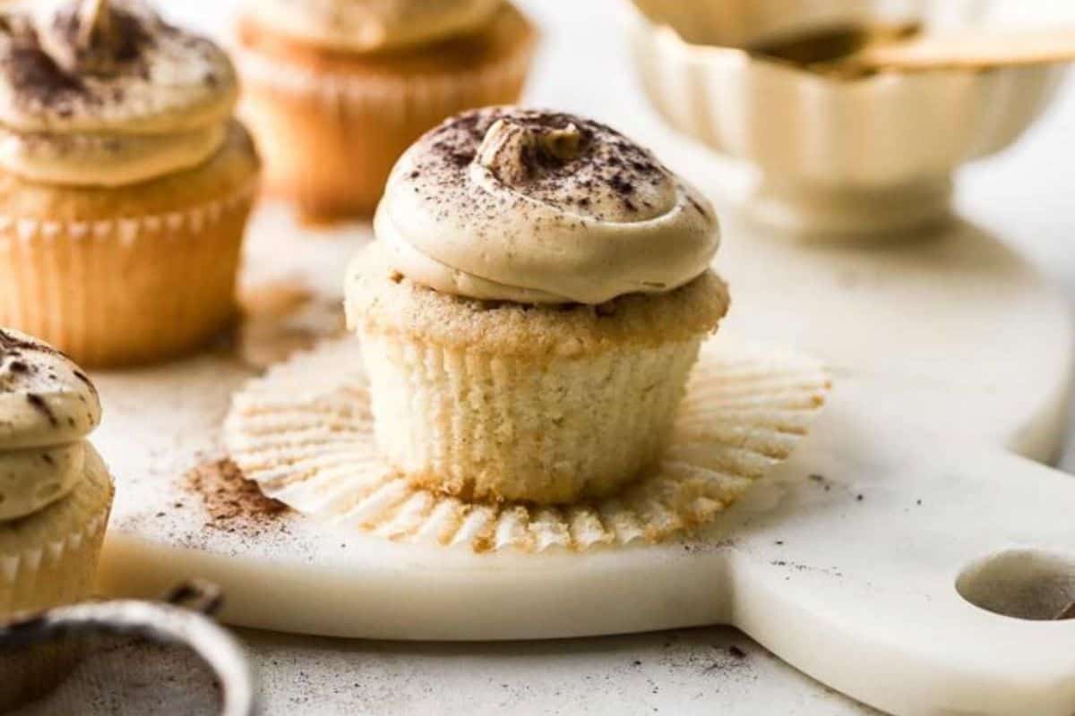 A close-up of vanilla cupcakes with swirled coffee-flavored frosting and a dusting of cocoa powder on top. The cupcakes sit on a light-colored marble cutting board, with one cupcake partially unwrapped. In the background, more cupcakes and a small bowl are visible.