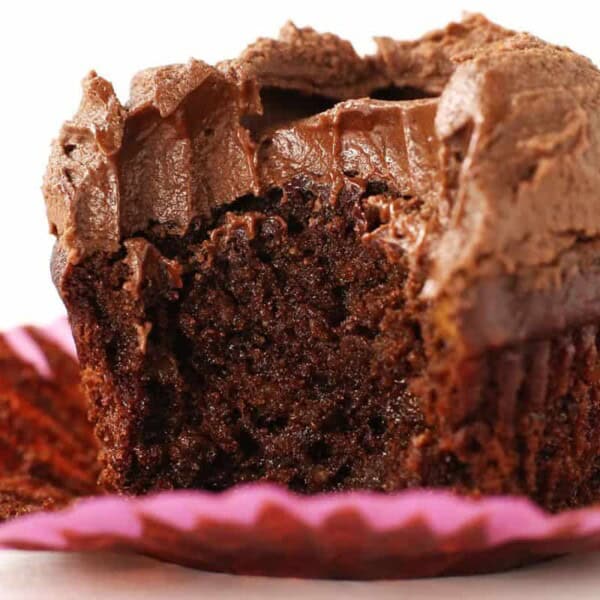 Close-up of a chocolate cupcake with a bite taken out, revealing a moist and rich interior. The cupcake is topped with a thick layer of creamy chocolate frosting and is placed in a pink cupcake liner.