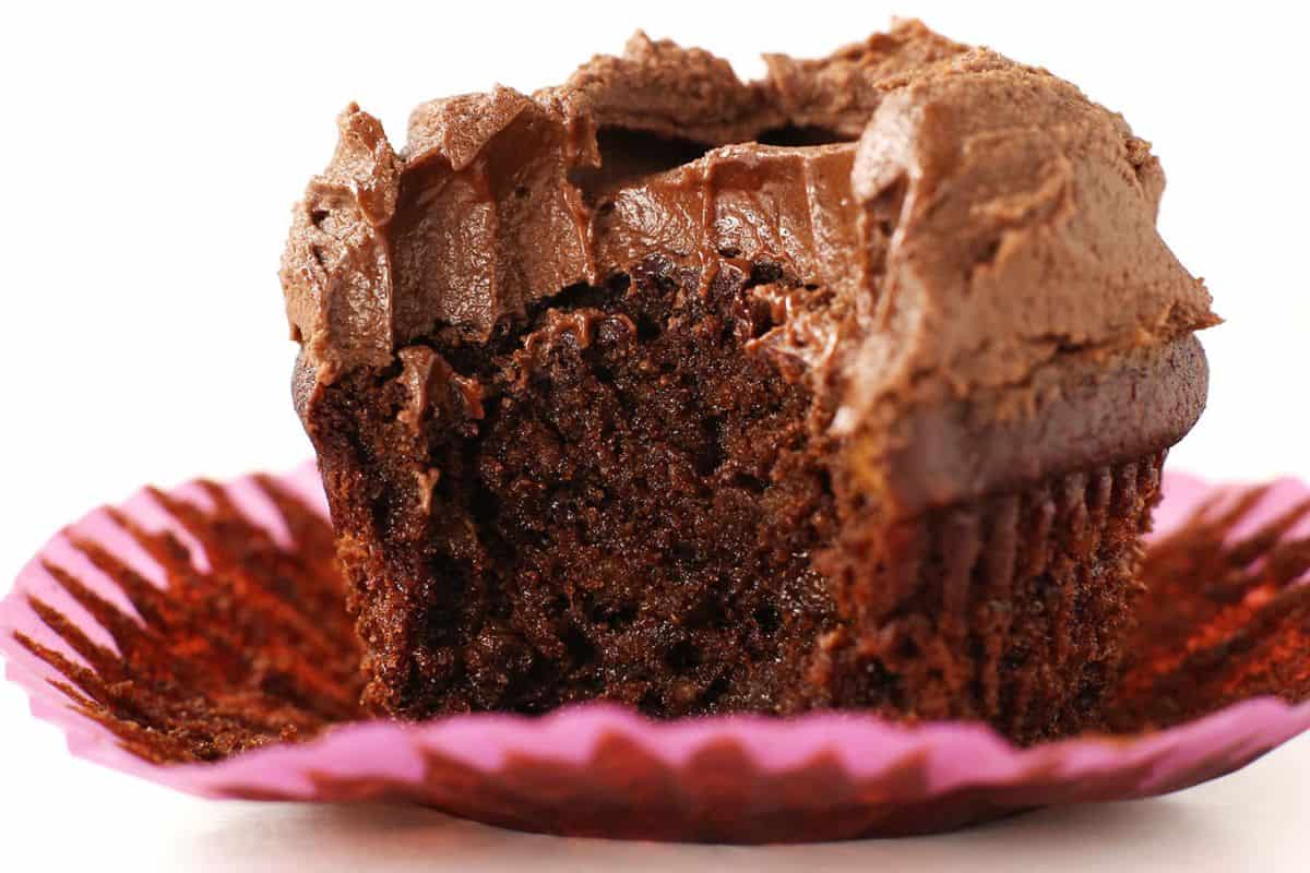 Close-up of a chocolate cupcake with a bite taken out, revealing a moist and rich interior. The cupcake is topped with a thick layer of creamy chocolate frosting and is placed in a pink cupcake liner.