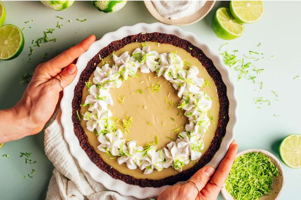 Two hands hold a white pie dish containing a key lime pie topped with whipped cream and lime zest. The pie sits on a light surface, surrounded by additional limes, a bowl of lime zest, and a white cloth.