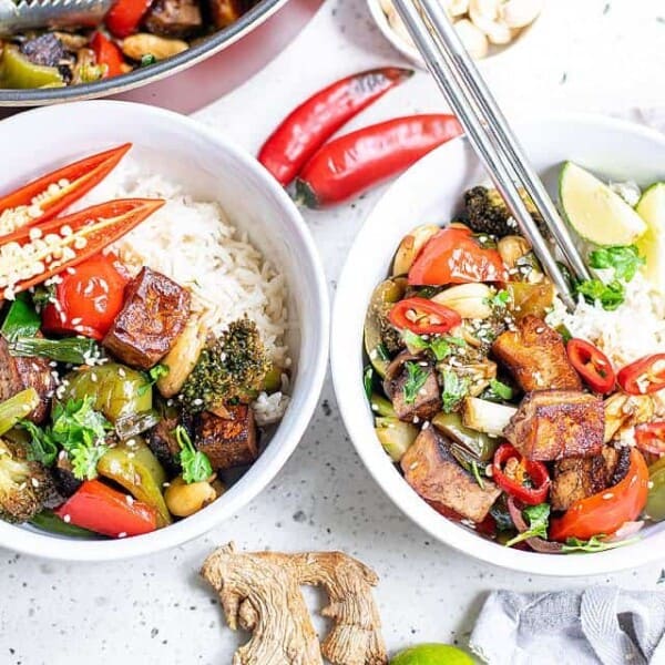 Two bowls of colorful rice and vegetable stir-fry are topped with various ingredients, including tofu, bell peppers, broccoli, and chili slices. Off to the side are ingredients like ginger, lime, and red chili peppers. Chopsticks rest on the bowls.