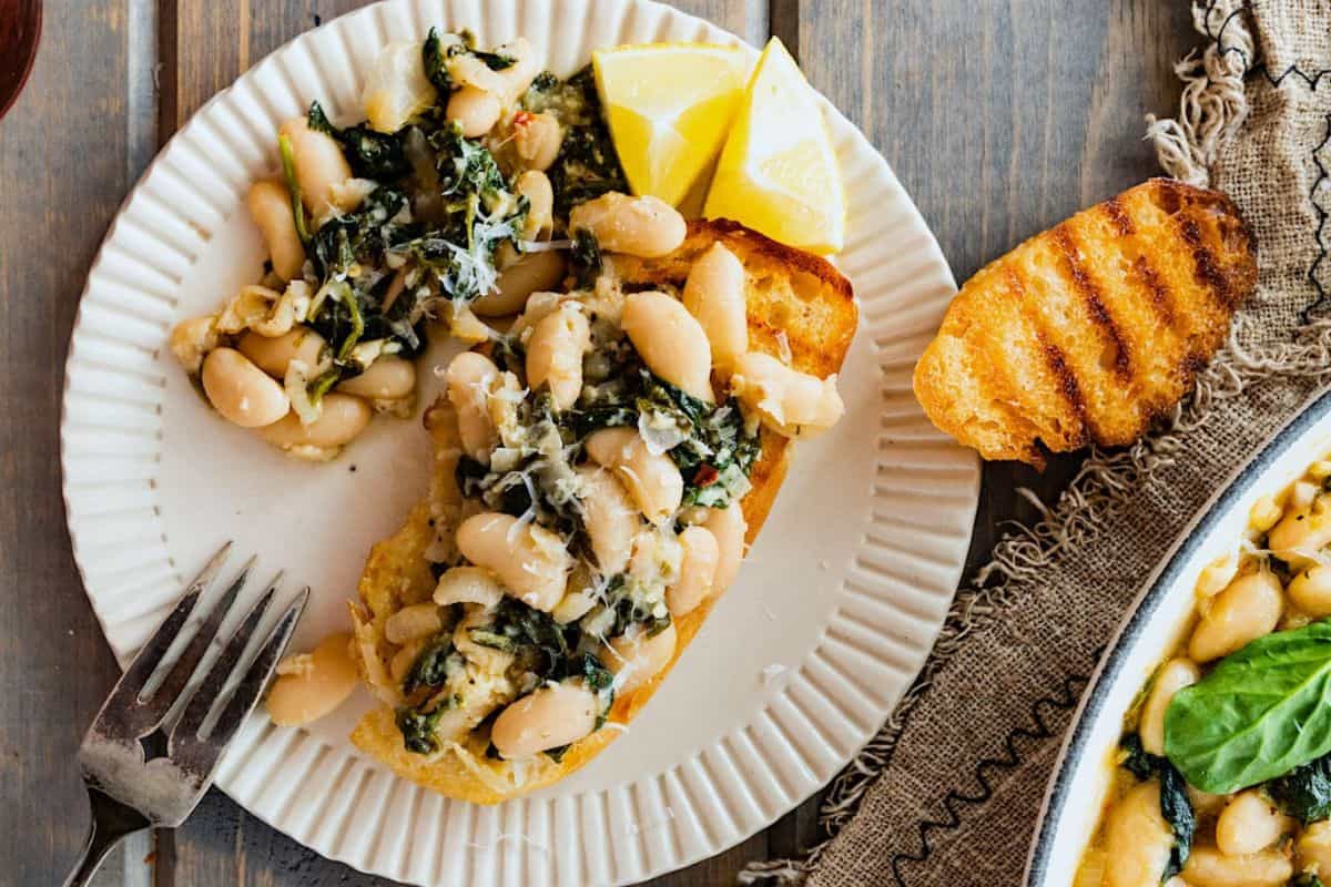 A white plate with a slice of toasted bread topped with a white bean, spinach, and herb mixture, garnished with grated cheese and two lemon wedges. A fork rests next to the plate, and another slice of toasted bread is partially visible on a burlap cloth.