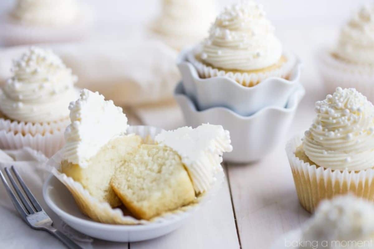 Close-up of vanilla cupcakes topped with white frosting and sprinkles. One cupcake is cut in half on a small white plate, revealing the fluffy texture inside. Another cupcake is placed in a white ruffled-edge container. A fork is set next to the plate.