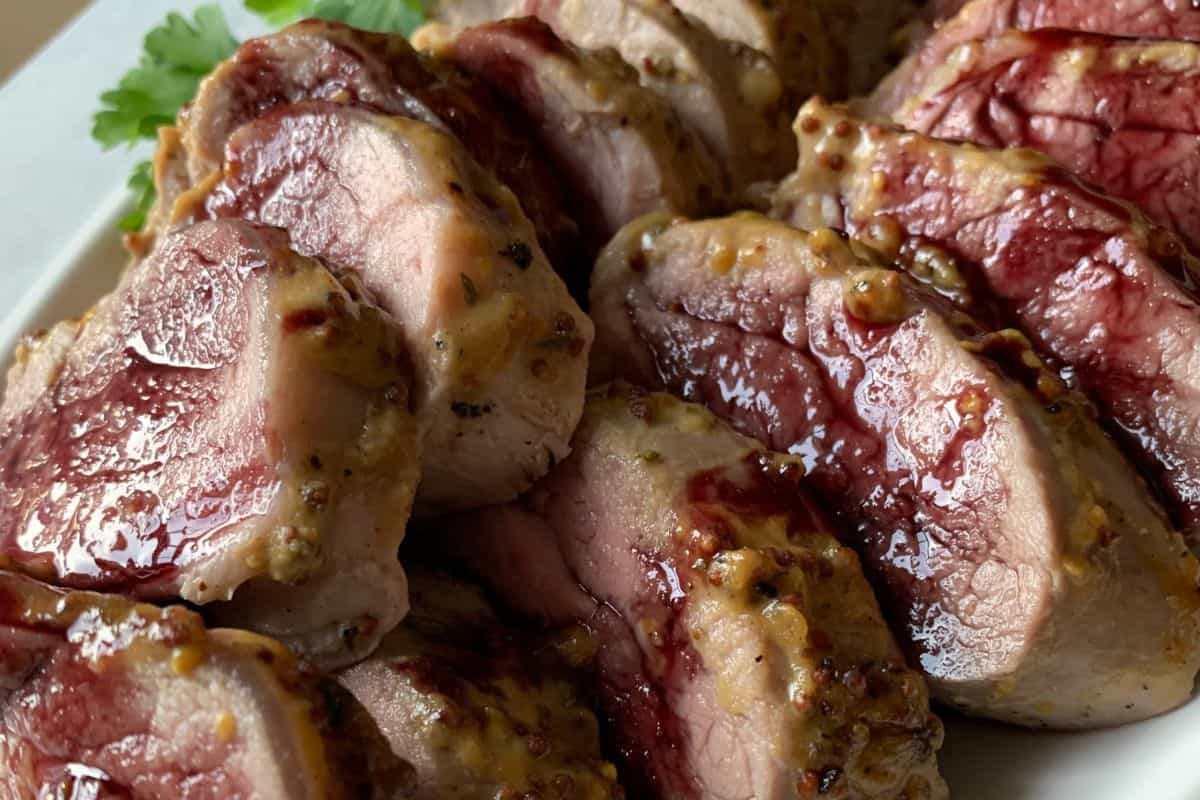 Close-up of a plate of sliced, medium-rare cooked beef tenderloin, garnished with a light herb seasoning. The slices reveal a slightly pink interior, indicating the medium-rare cooking style. Parsley leaves are visible in the background for garnish.