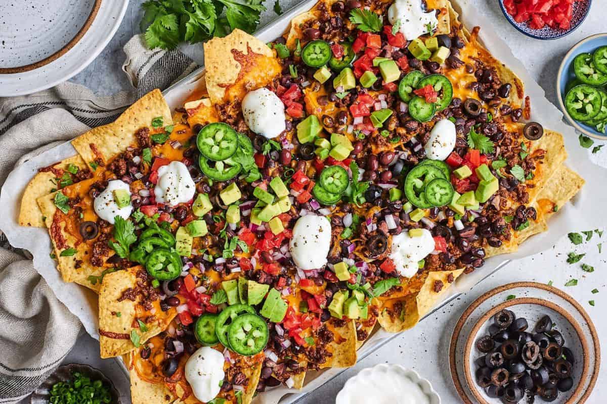 A platter of loaded nachos topped with melted cheese, seasoned ground beef, sliced jalapeños, diced tomatoes, black olives, avocado chunks, red onions, sour cream dollops, and fresh cilantro. Surrounding the platter are bowls of additional diced tomatoes, black olives, and sour cream.