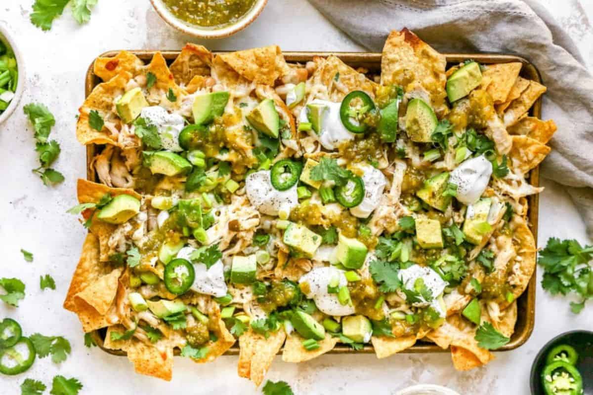 An overhead view of a tray of loaded nachos topped with melted cheese, avocado chunks, sliced jalapenos, dollops of sour cream, green salsa, and fresh cilantro. There are bowls of green salsa and cilantro garnishes around the tray.